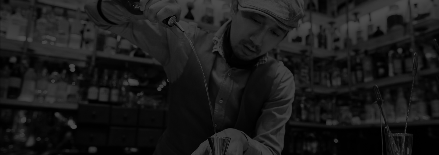 Darkened black and white image of bartender free pouring alcohol in
        to a jigger